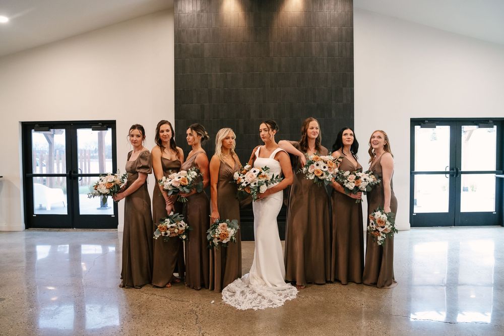 Bride in white lace gown poses with bridesmaids in brown dresses holding floral bouquets in elegant venue.