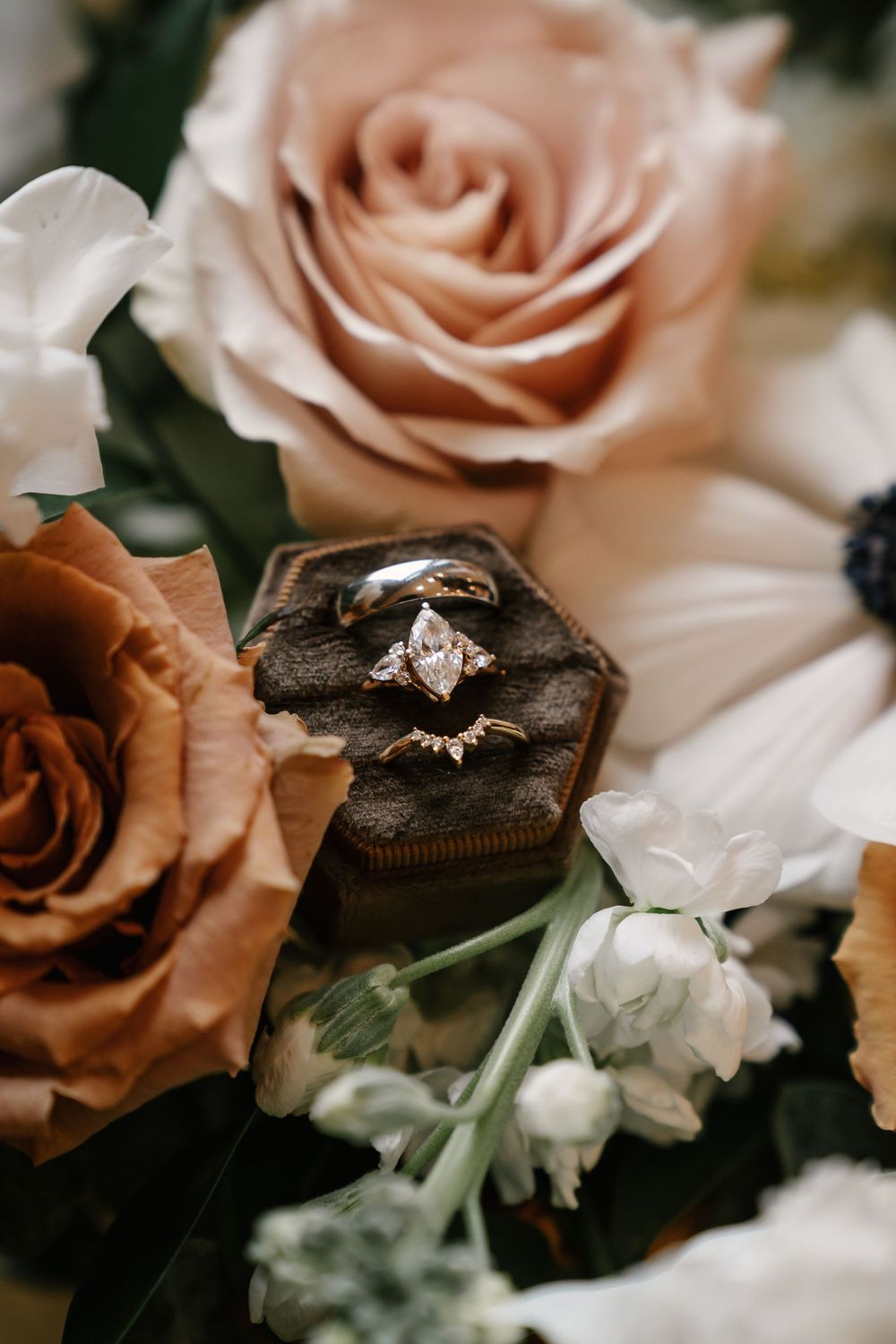 Gold engagement ring with marquise diamond in velvet box surrounded by blush, rust, and white roses.
