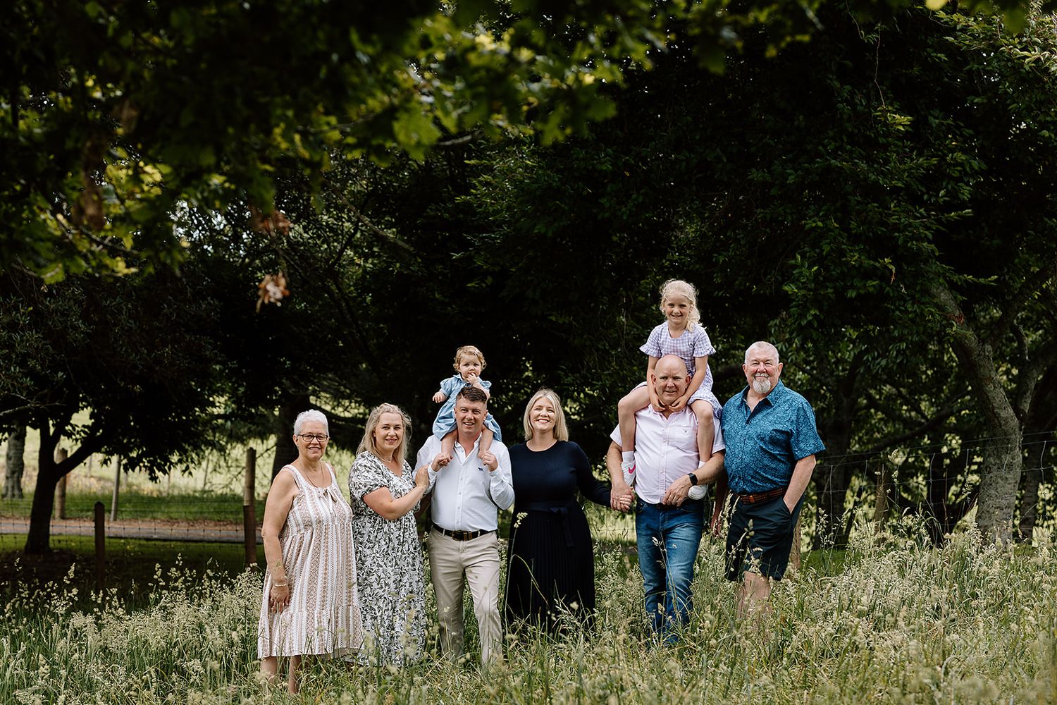A multi-generational group standing together outdoors in a meadow with tall grass and trees in the background.