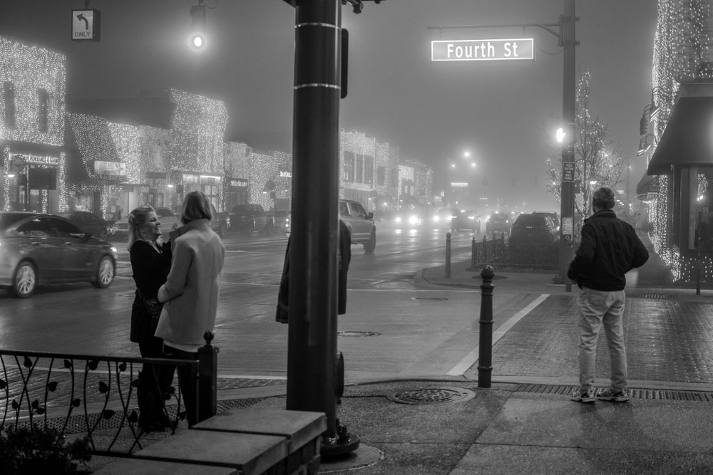 Foggy night street scene with silhouettes of people and glowing storefronts in black and white.
