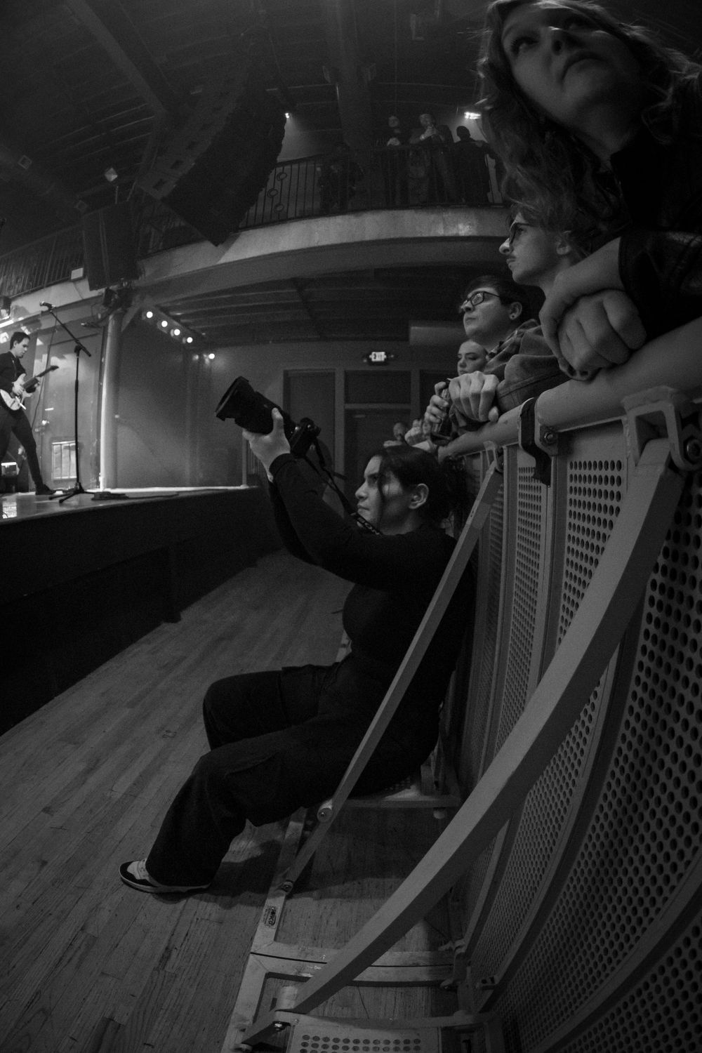 Black and white photo of concert photographer shooting from stairs while audience watches live performance.