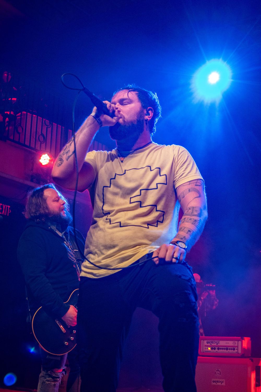 Tattooed vocalist performs on stage with microphone under blue stage lights at a live rock concert.
