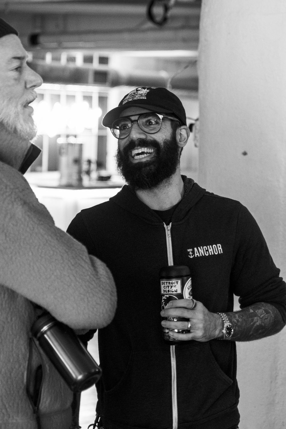 Bearded man in Anchor hoodie and cap smiles while holding a drink at a casual social gathering.