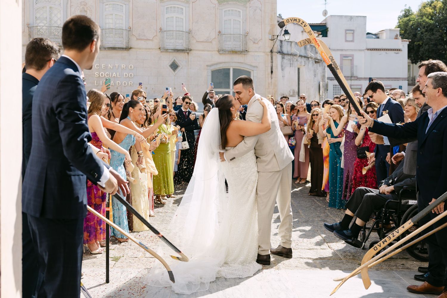 Bride and groom share a kiss during wedding ceremony exit while guests form an arch with gold oars outdoors.