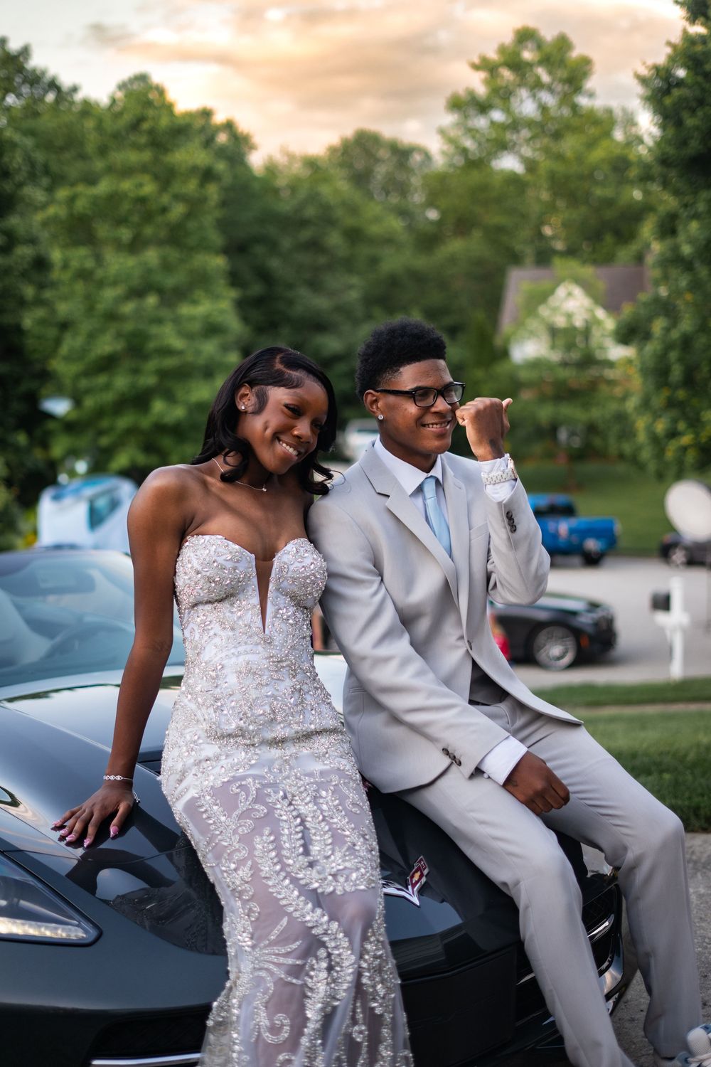A young couple in prom attire poses on a black car, wearing a beaded gown and gray suit at sunset.