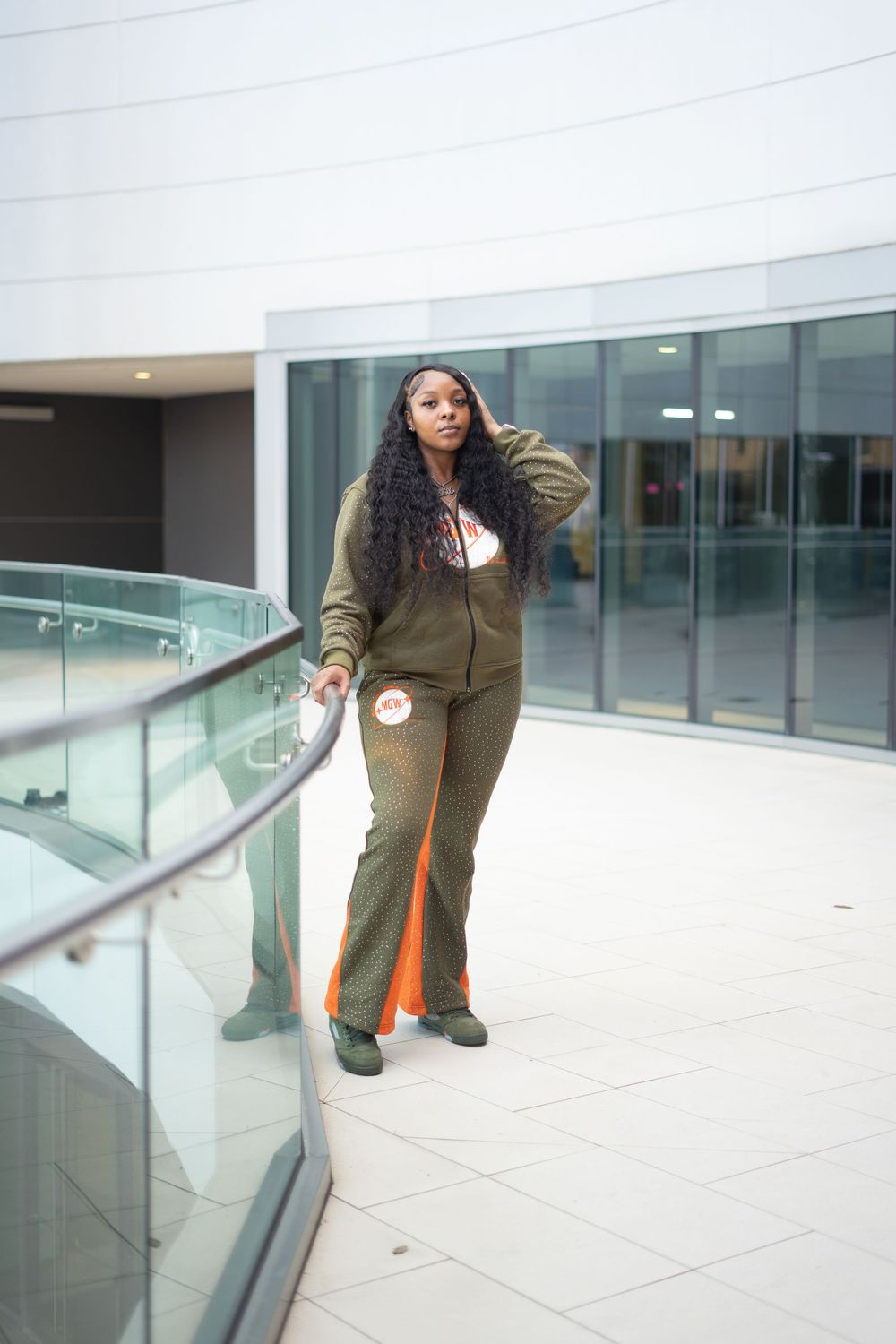 Woman in olive green tracksuit with orange stripe poses by glass railing in modern building interior.