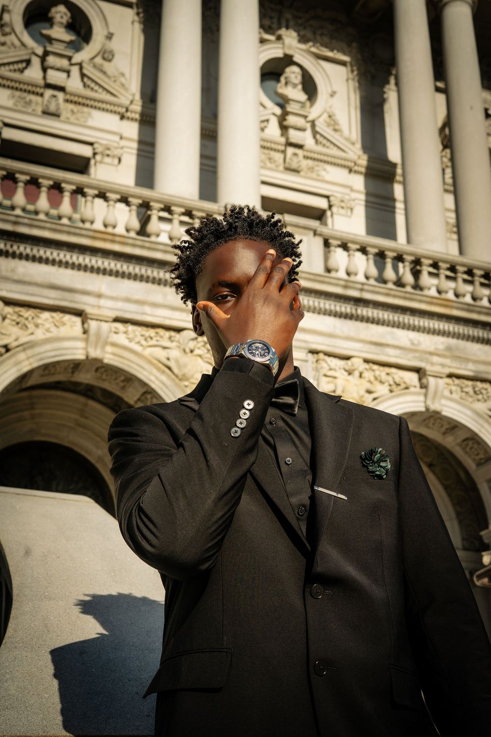 Stylish Black man in dark suit with green pocket square and luxury watch poses before ornate classical building facade.