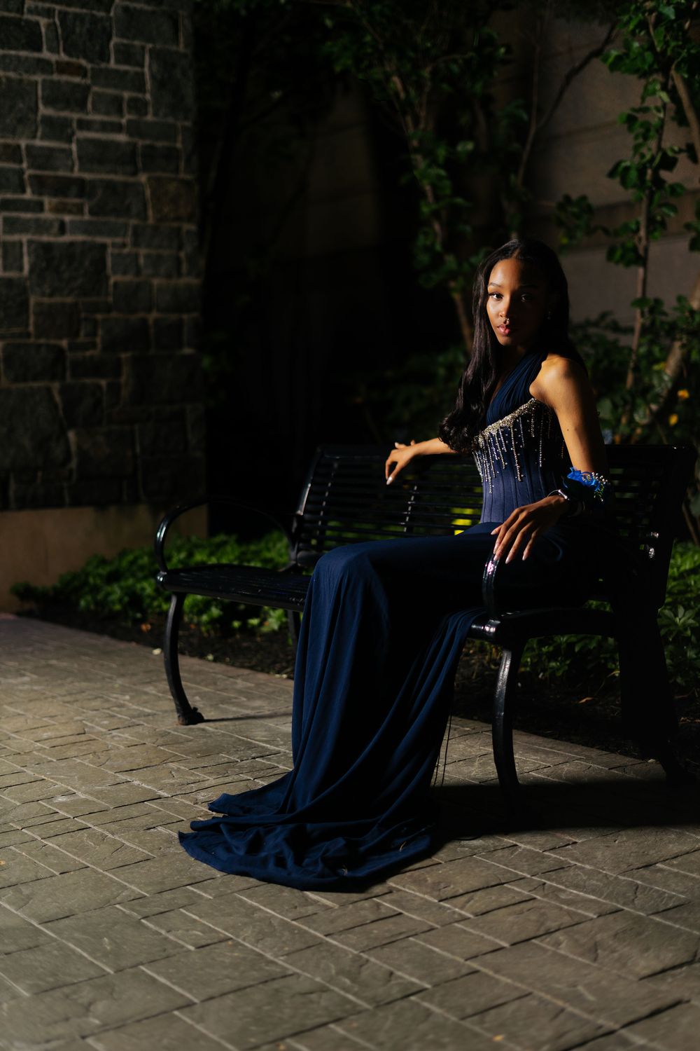Young woman in elegant navy blue gown seated on a bench at night, posing against a stone wall with ivy.