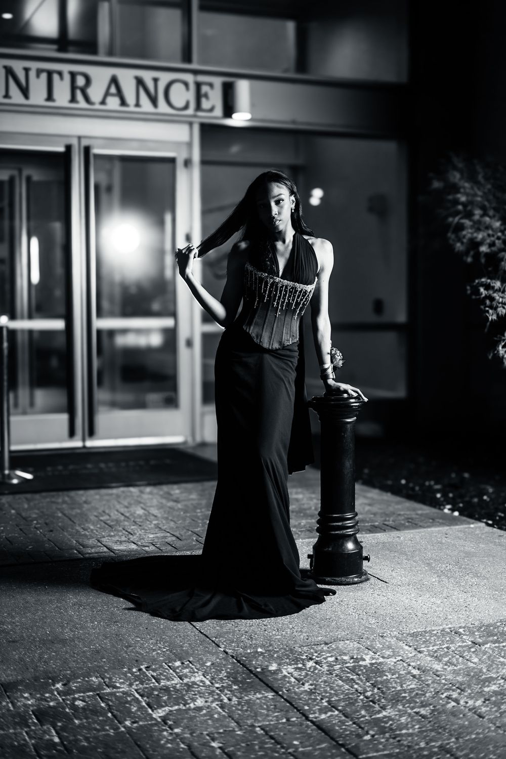 Black and white photo of elegant woman in long black gown posing by lamppost outside building entrance at night.