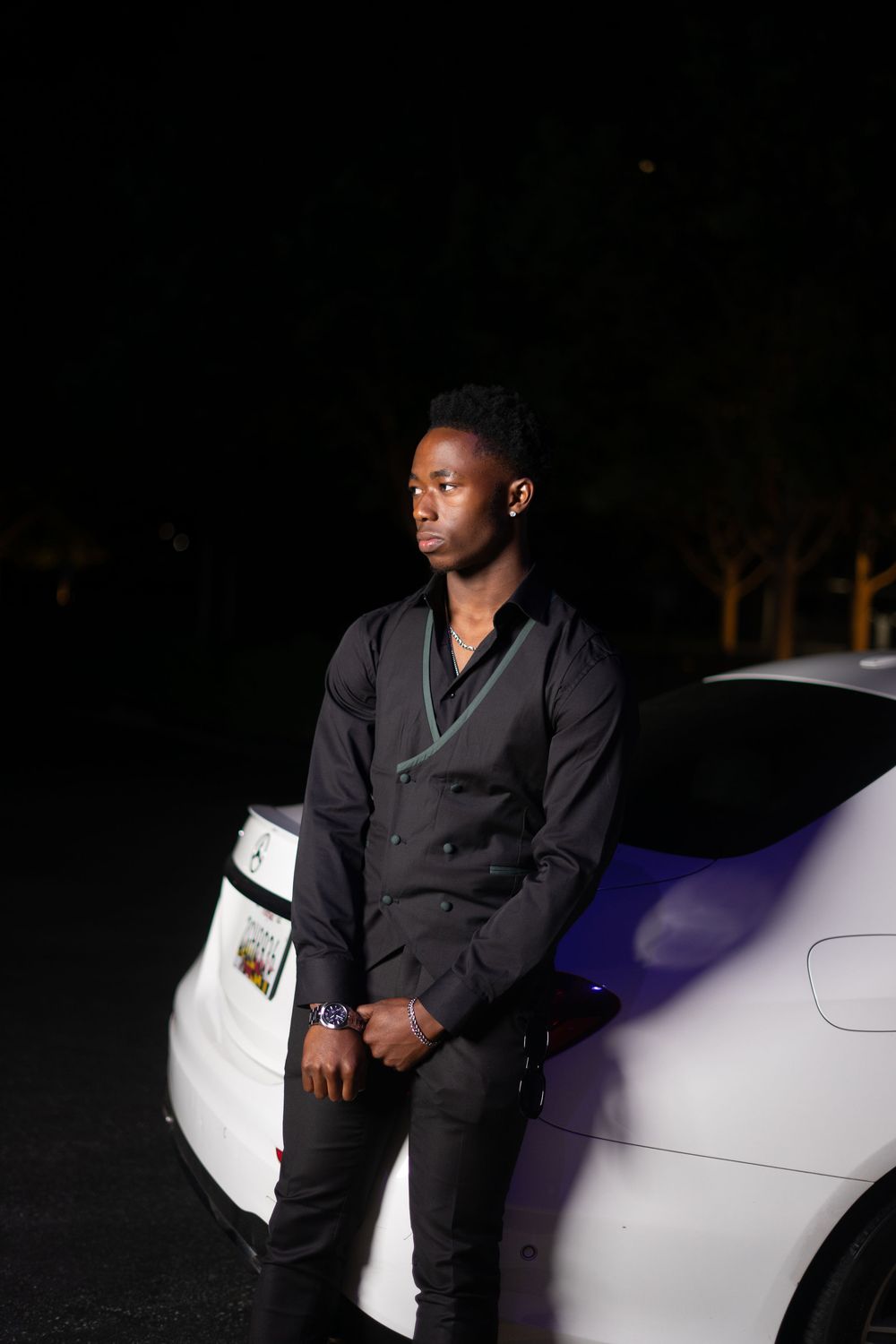 Young Black man in black shirt leans against white car at night, looking away confidently.
