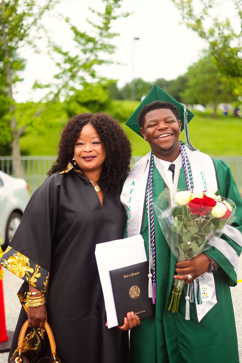 A proud graduate in green cap and gown holds roses beside a woman in black robe holding a diploma outdoors.