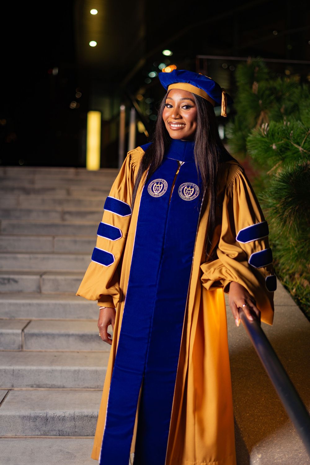 Woman in blue and gold doctoral graduation regalia smiles on steps at night.