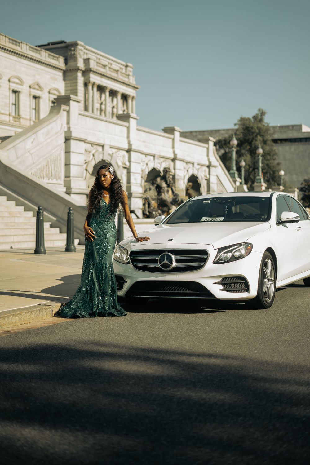 Woman in green sequin gown posing beside white Mercedes-Benz sedan near grand building with stone staircase.