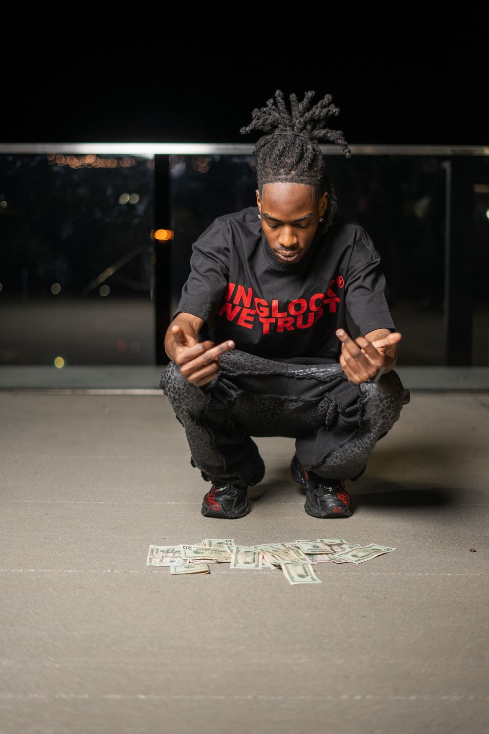 Young man with dreadlocks crouching over scattered dollar bills on pavement, wearing black and red graphic hoodie.