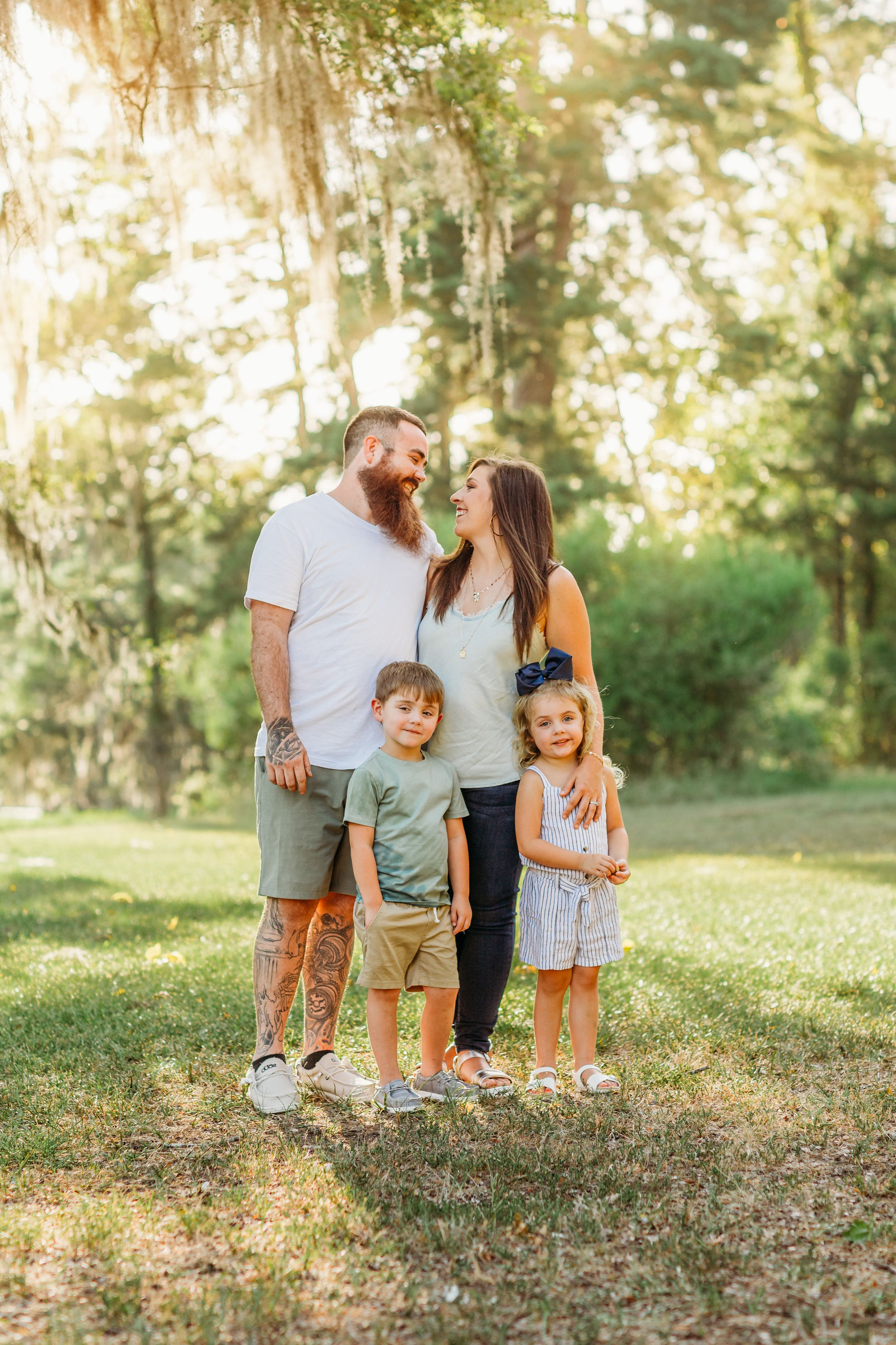 Loving family of four standing together outdoors in a sunlit wooded area on a warm summer evening.