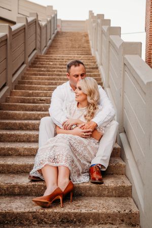 A couple in white clothing sits romantically on concrete stairs at sunset, with her wearing red heels and a lace dress.