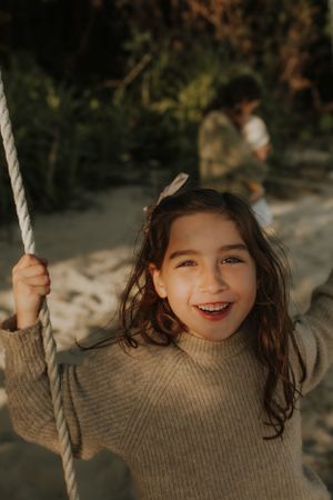 Person wearing a beige sweater smiles while holding a rope swing in an outdoor setting during golden hour.