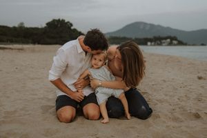 A family shares a tender moment together while sitting on a sandy beach with mountains visible in the background.