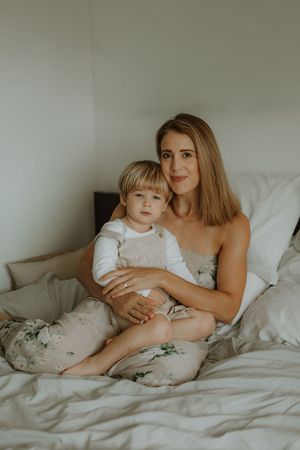 Parent and child share tender moment while relaxing together on white bedding in soft natural light.