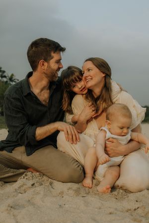 A loving couple and their baby share intimate moments while laying on a sandy beach at sunset.