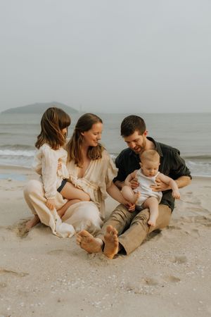 A family in white and neutral clothing sits together playfully on a misty beach.