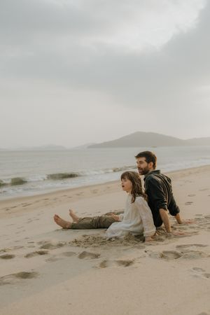 Two people laying side by side on a sandy beach with mountains and cloudy skies in the background.