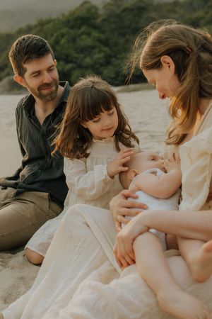 A family sits together on a beach at sunset, sharing a tender moment with their newborn baby.