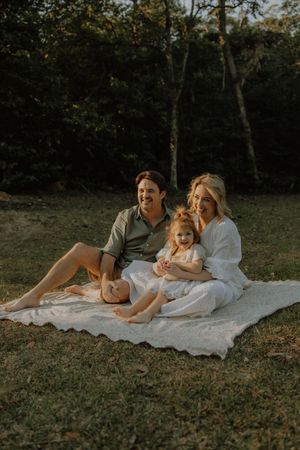 A family relaxes together on a white blanket during a golden hour photoshoot in a wooded setting.