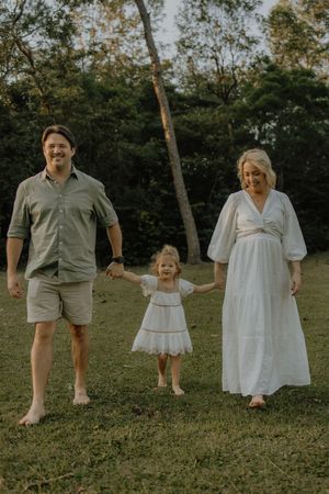 A family in coordinating white and beige outfits walks together holding hands in a grassy outdoor setting at sunset.