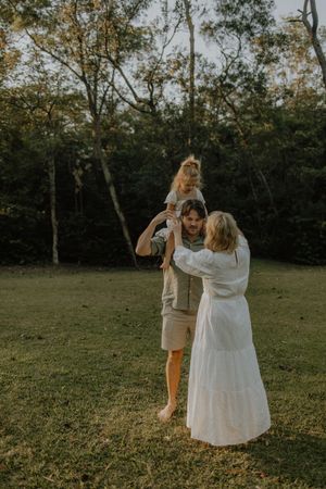 A family shares a tender moment together in a sunlit grassy field surrounded by trees at golden hour.