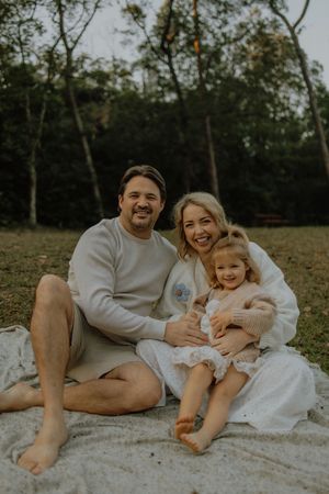 A family relaxes together on a white blanket during an outdoor photoshoot in a wooded setting.