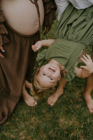 A laughing child hangs upside down between a pregnant woman and another person in green dress outdoors.