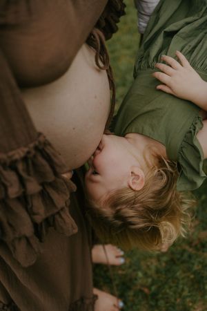 A young child kisses a pregnant woman's bare belly outdoors, both wearing earthy toned clothing on green grass.