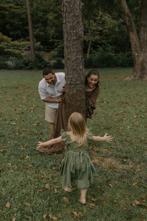 A joyful family plays peek-a-boo around a tree in a lush green park, with a toddler in a green dress running toward her parents.