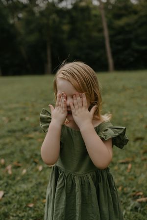 Young girl in green ruffle dress covering her face with hands while standing on grass in a park.