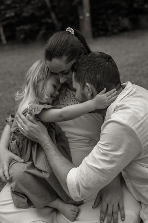 Black and white photo of parents embracing their young blonde daughter in a tender family moment outdoors.