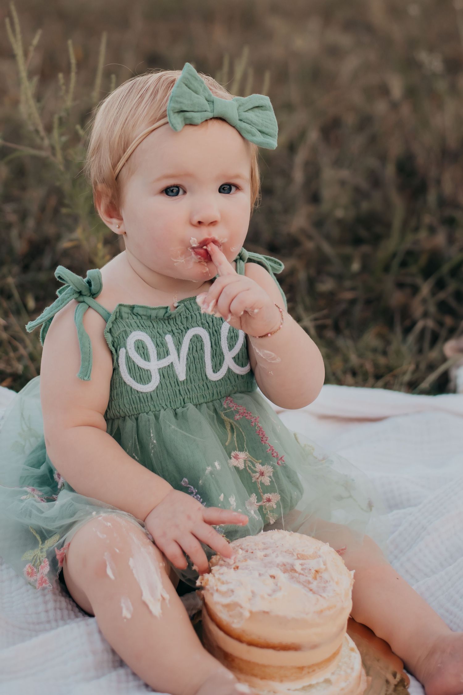 Baby in mint green dress sits on blanket with birthday cake and matching bow.