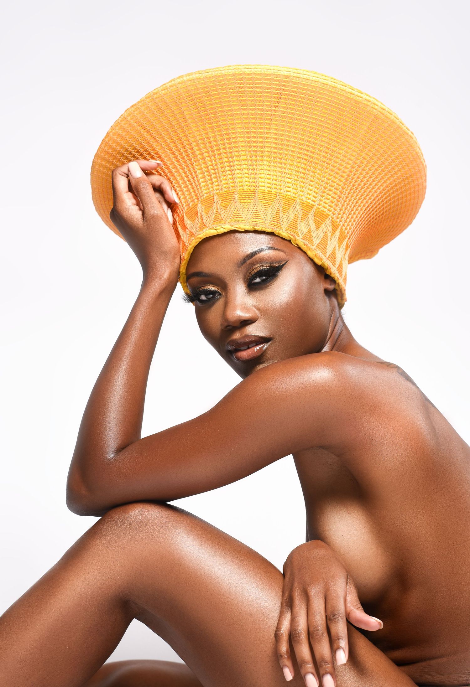 A Black woman poses gracefully wearing a bold orange woven hat with geometric patterns against a white background.