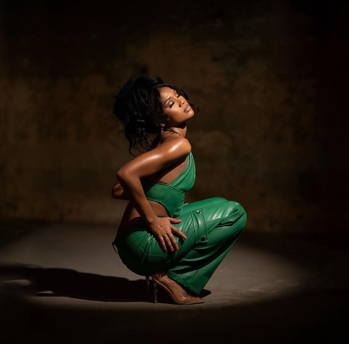 A woman in a green leather jumpsuit poses gracefully on the floor in dramatic studio lighting.