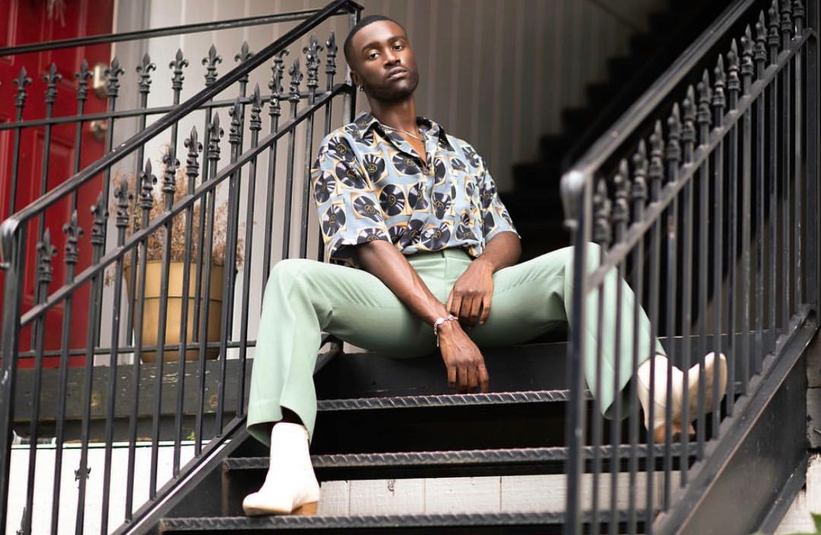 Man sitting on staircase wearing floral print shirt and mint green pants with white shoes.