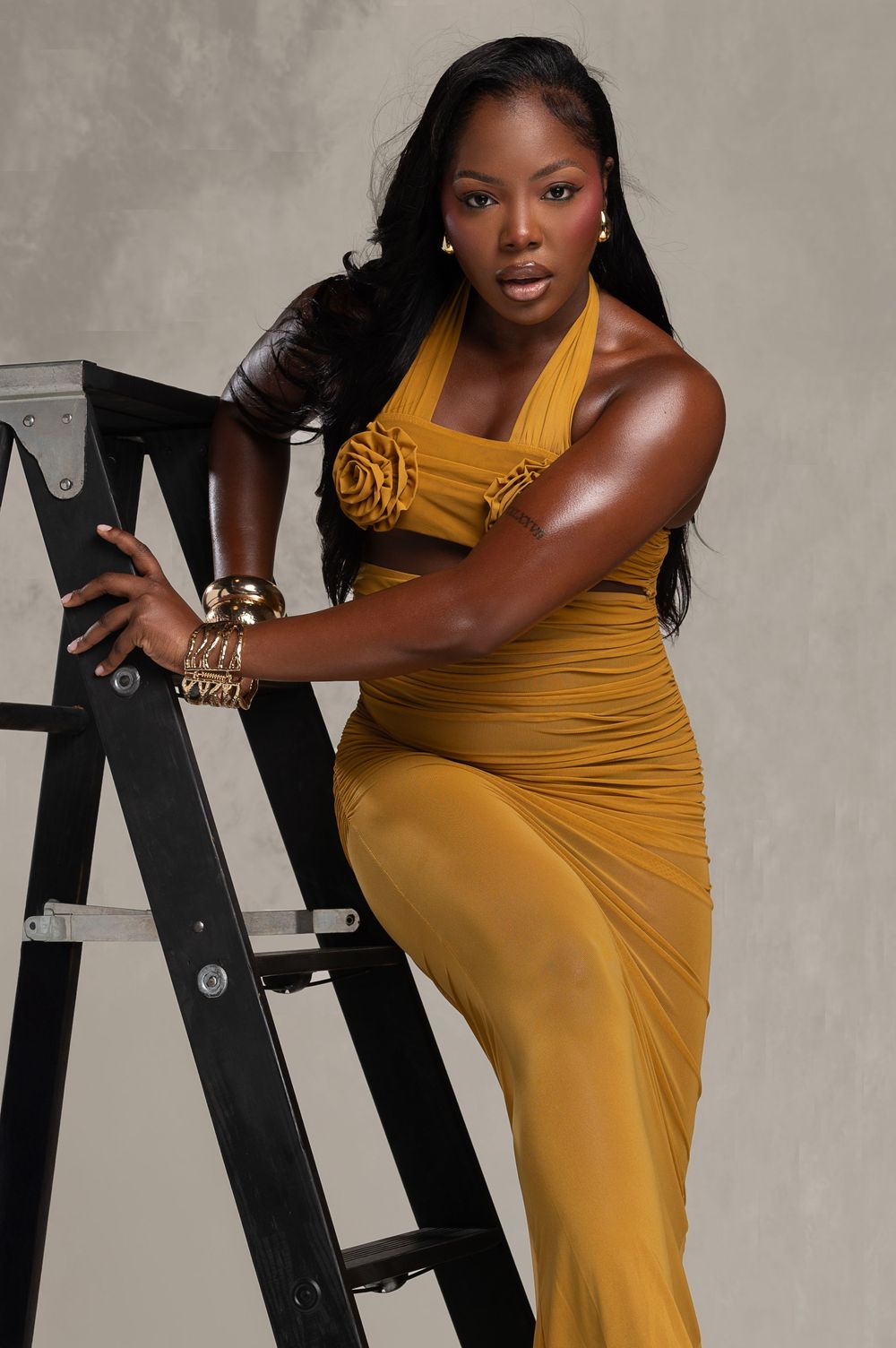 Woman in mustard yellow halter gown poses elegantly on black ladder with gold accessories and long black hair.