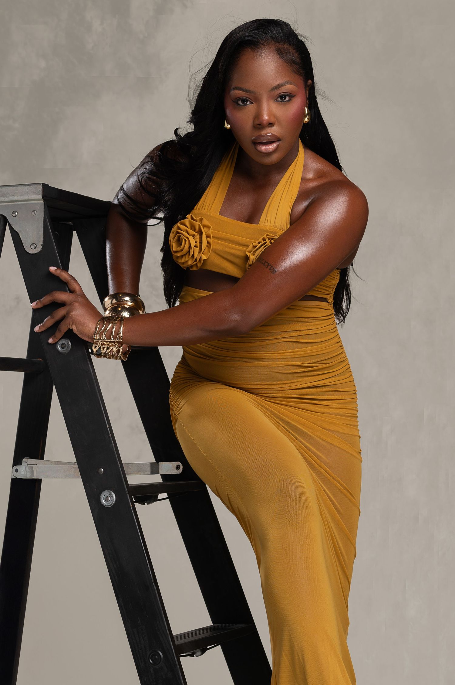 Woman in mustard yellow halter gown poses elegantly on black ladder with gold accessories and long black hair.