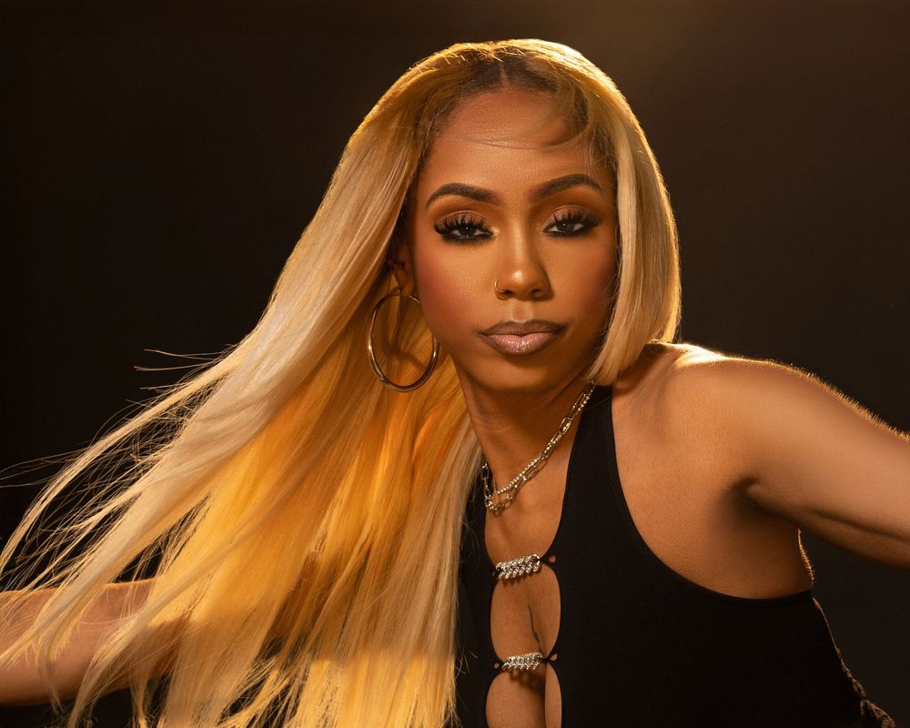 Woman with long blonde hair wearing a black outfit posing confidently against a dark background.