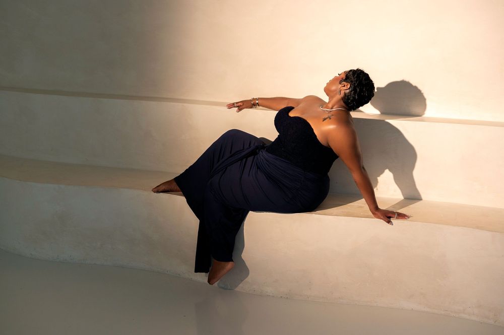 Woman in navy blue strapless jumpsuit reclines gracefully against a white wall, casting dramatic shadows.