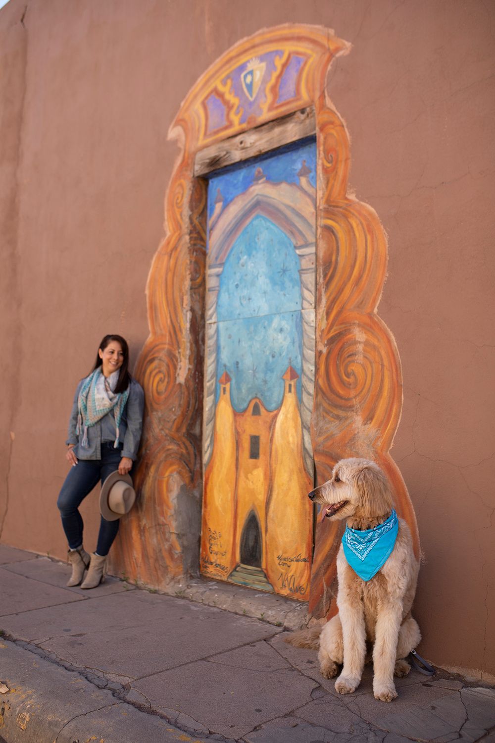 A dog and companion pose in front of a colorful southwestern mural painted on an adobe wall in Santa Fe, New Mexico.