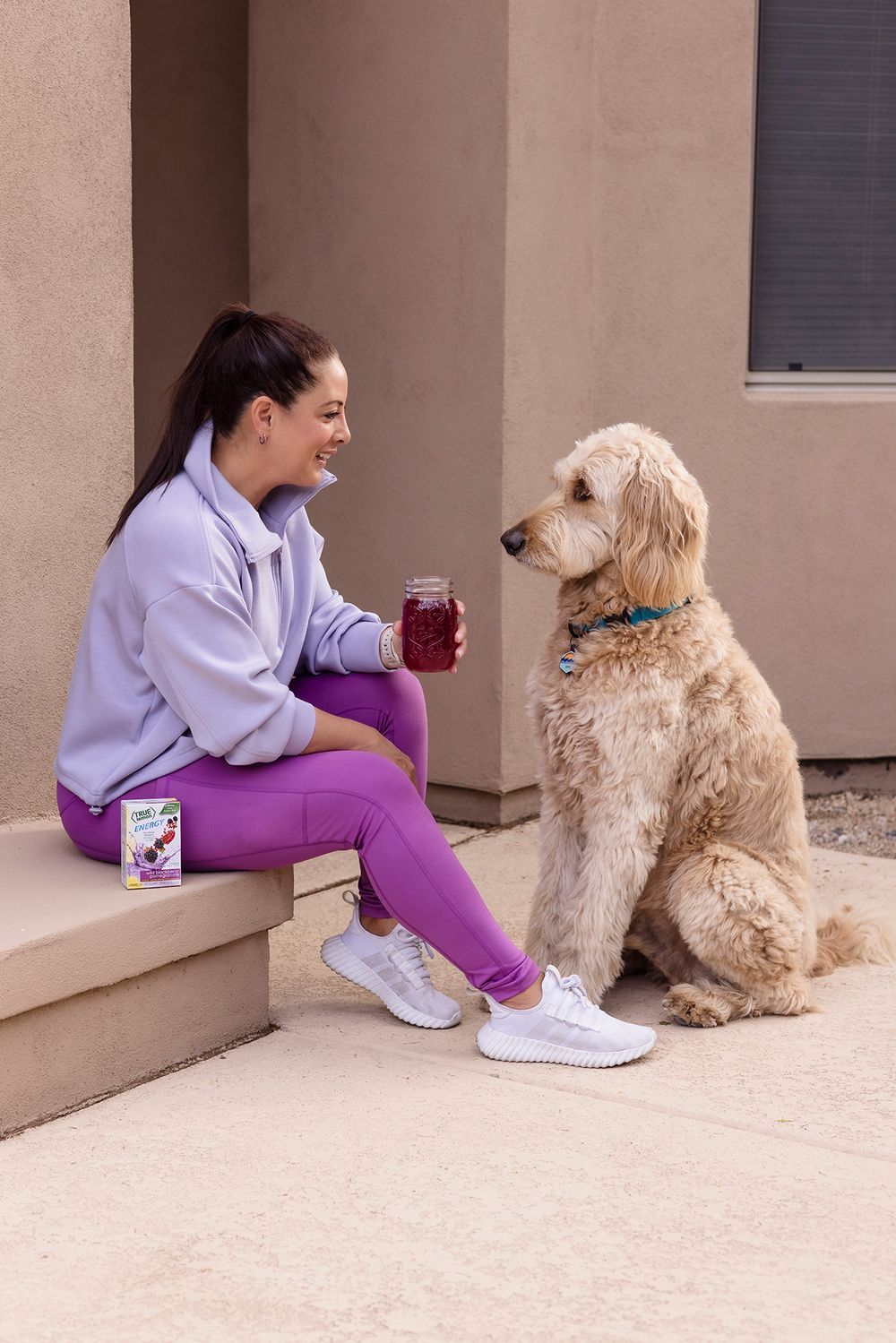 A person in purple workout clothes sits on steps with their goldendoodle dog and a beverage.