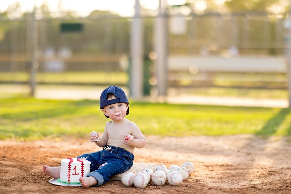 Baby boy in baseball cap sits on dirt field with cake and baseballs for his first birthday photo.