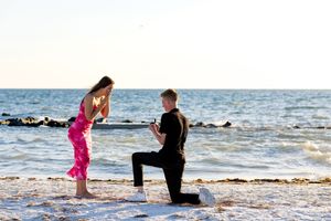 Man proposes on a beach at sunset, kneeling before a woman in a pink outfit near the ocean.