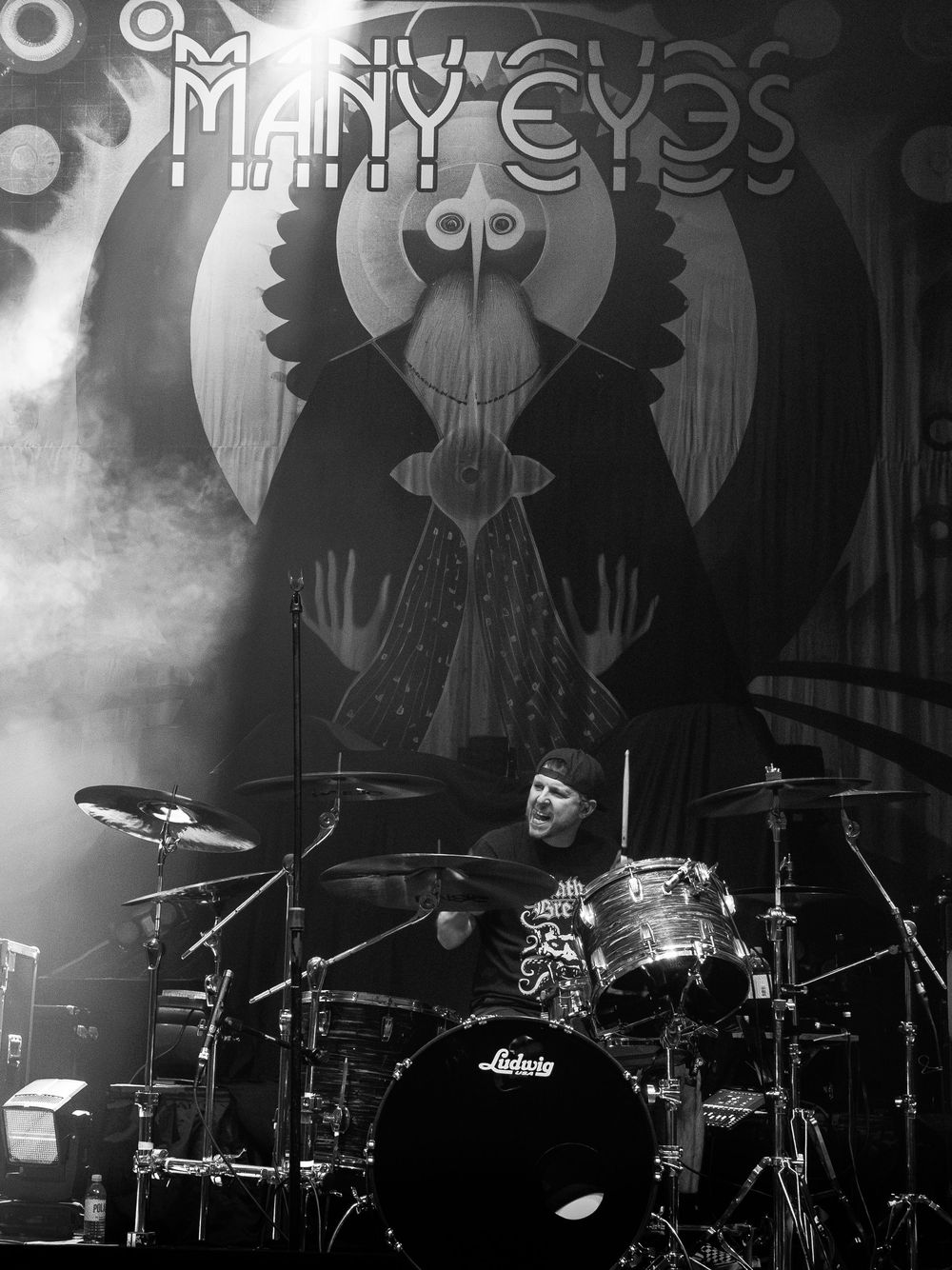 Black and white photo of a drummer performing on stage behind a Ludwig drum kit with a Many Eyes banner and stage smoke.
