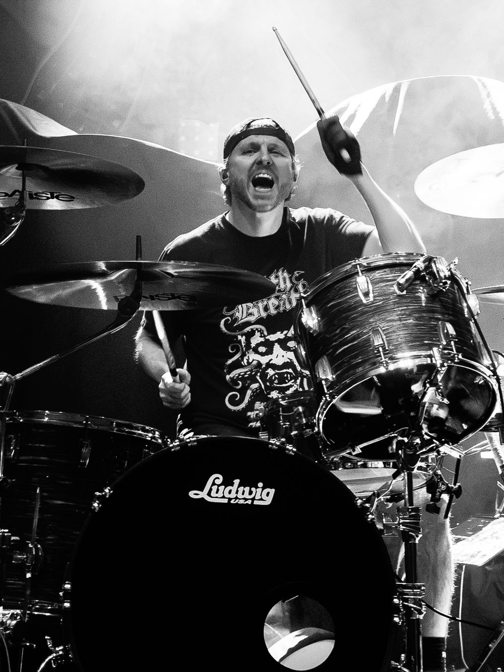 Black and white photo of an energetic drummer in a backwards cap and graphic tee playing a Ludwig drum kit with cymbals on stage.